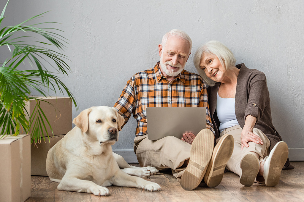 Older woman and man sitting on floow with dog looking at a computer surrounded by moving boxes