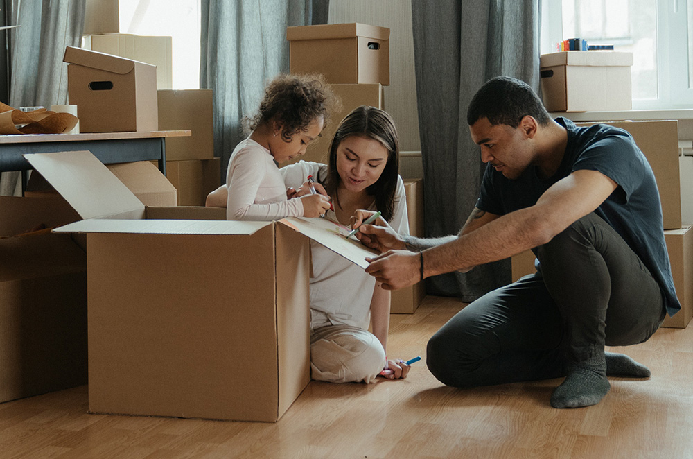 Family surrounded by moving boxes with small girl inside a box writing on the flap