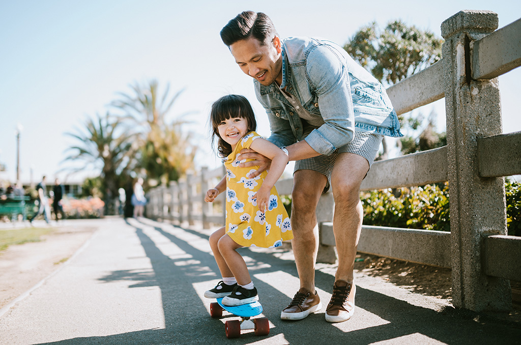 Dad and daughter riding on a skateboard outside.