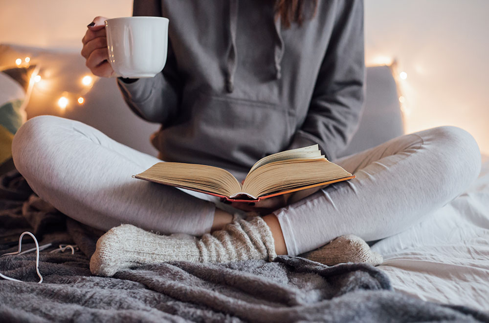 Woman reading book with cup in hand in bed