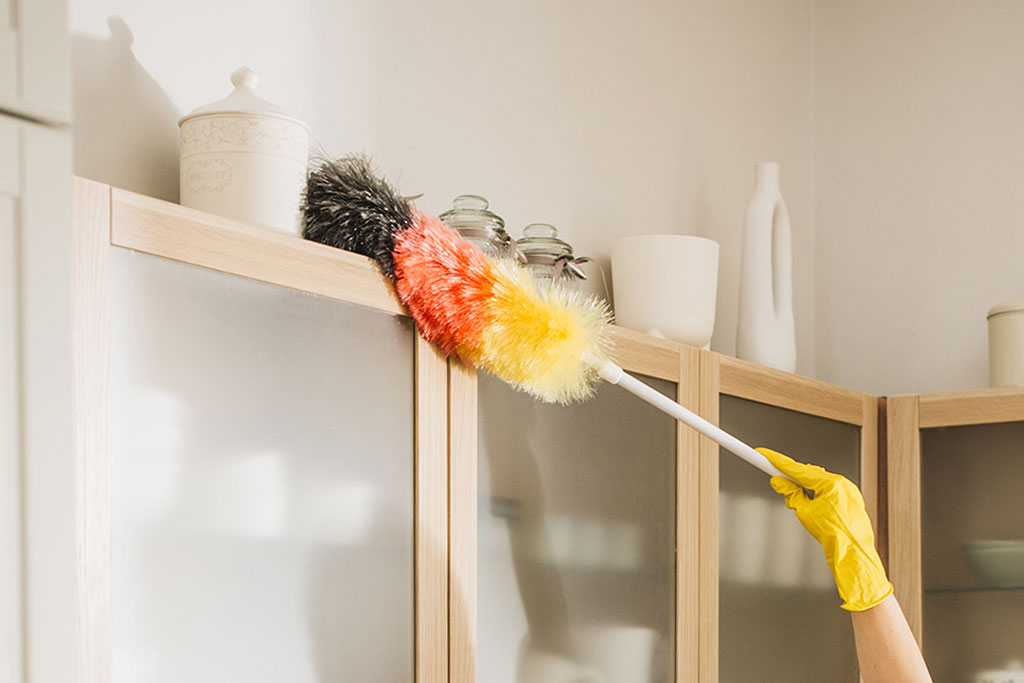 Dusting top shelves of cabinet in kitchen